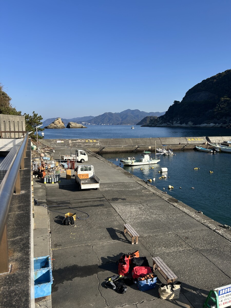 快晴の雲見港と静かな海、ダイビング器材が並ぶ朝の準備風景。前方には牛着岩が見える穏やかな港の様子。